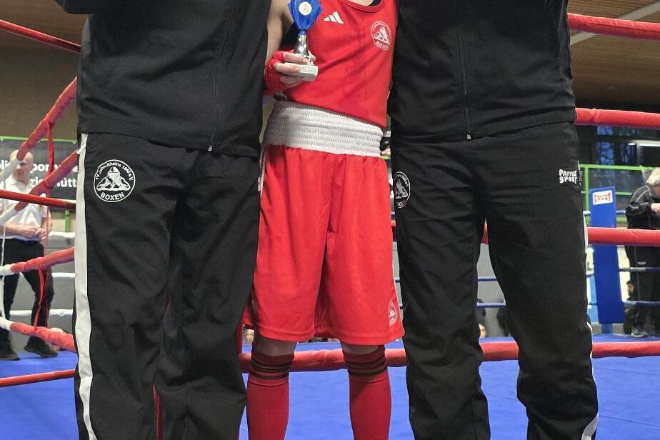 Drei Personen stehen in einem Boxring; ein Mädchen in roter Boxkleidung hält einen Pokal, flankiert von zwei Trainern.