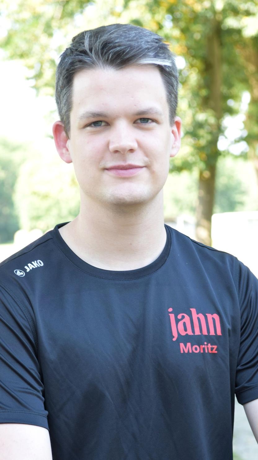 Junger Mann mit kurzem, grauem Haar, tr&auml;gt ein schwarzes Sportshirt mit rotem Aufdruck, sitzt im Freien unter B&auml;umen.