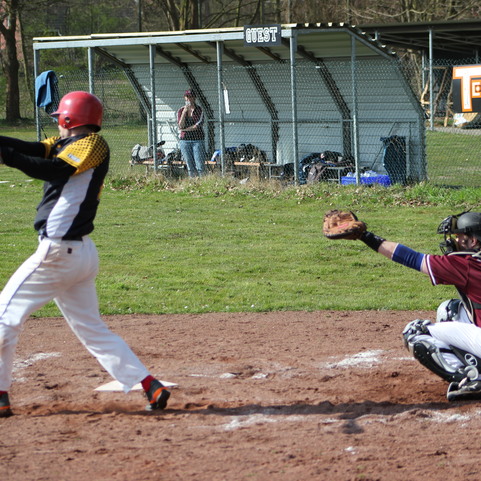 Baseballspieler beim Schlagversuch, w&auml;hrend der Catcher bereit ist und Zuschauer im Hintergrund stehen.
