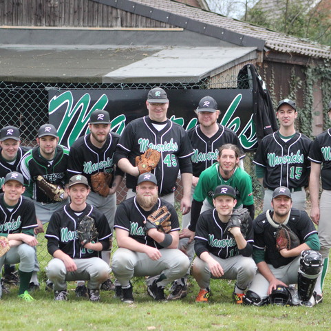 Gruppe von 15 Baseballspielern in schwarz-gr&uuml;nen Trikots, posierend vor einem Zaun mit Vereinslogo und einem Holzgeb&auml;ude.