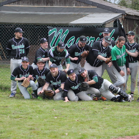 Gruppenfoto mit einem Baseballteam in schwarz-gr&uuml;nen Trikots, l&auml;chelnd vor einem Zaun und einer Beschriftung im Hintergrund.
