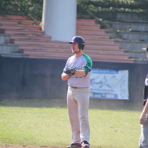 Baseballspieler steht an der Base, w&auml;hrend ein Schiedsrichter in Uniform daneben beobachtet. Stadionbleacher im Hintergrund.