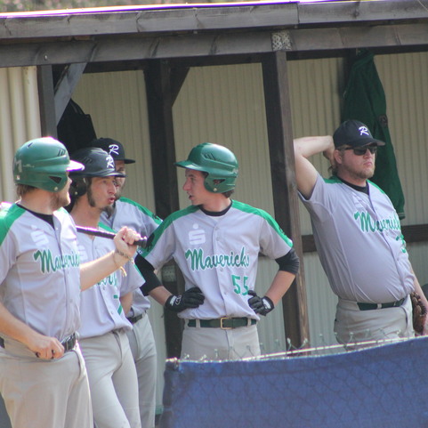 Vier Baseballspieler in Uniform stehen an der Bank, einer h&auml;lt einen Schl&auml;ger, w&auml;hrend die anderen mit ihm sprechen.