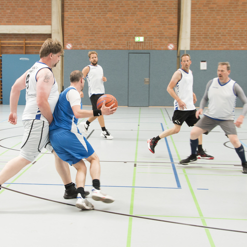 Sechs M&auml;nner spielen Basketball in einer Sporthalle, w&auml;hrend ein Spieler mit Ball einen anderen dribbelt.