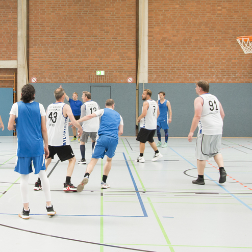 Zwei Basketballmannschaften spielen in einer Halle, ein Spieler wirft den Ball zum Korb.