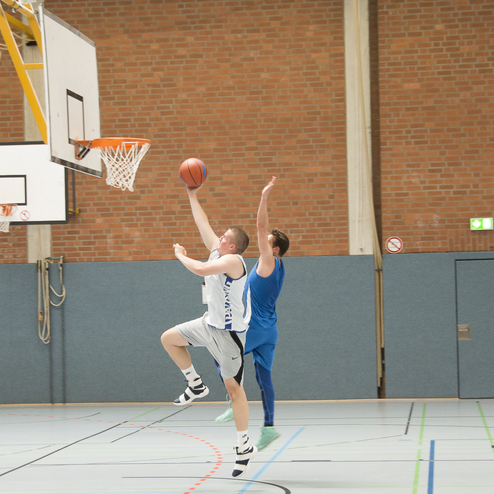 Basketballspieler hebt ab zum Korb, w&auml;hrend ein Gegner versucht, ihn zu blocken. Sporthalle mit Basketballkorb im Hintergrund.