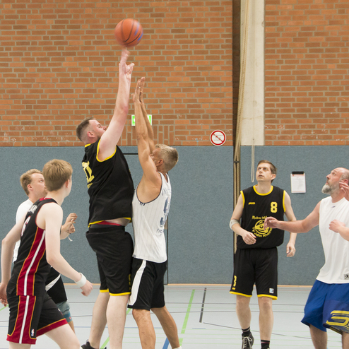 Zwei Basketballspieler springen zum Ballwurf in einem Sporthallen-Setting, mehrere Spieler im Hintergrund.