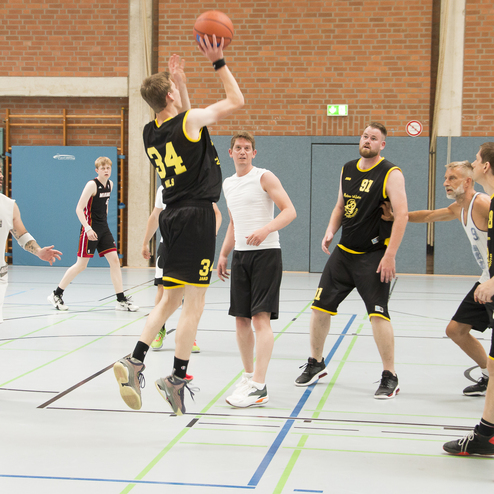 Zwei Basketballteams spielen in einer Sporthalle; ein Spieler springt zum Wurf mit dem Ball in der Hand.