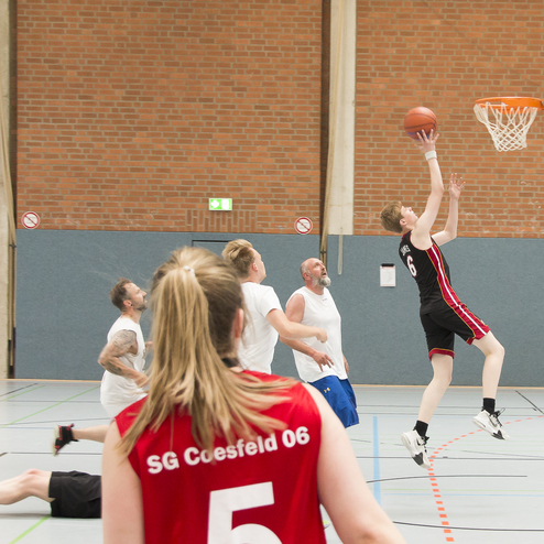 Basketballspieler macht einen Sprungwurf in einer Halle, w&auml;hrend andere Spieler im Hintergrund beobachten.