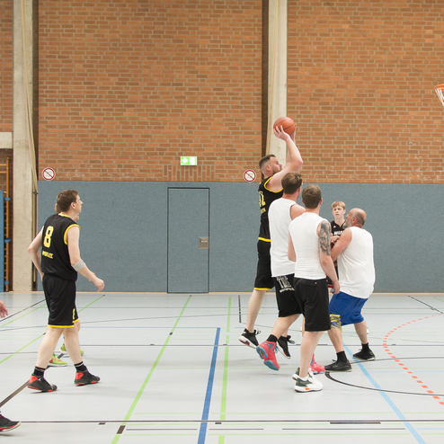 Basketballspieler springen zum Korb, w&auml;hrend andere Verteidiger versuchen, den Wurf zu blocken. Halle mit Holzfu&szlig;boden.
