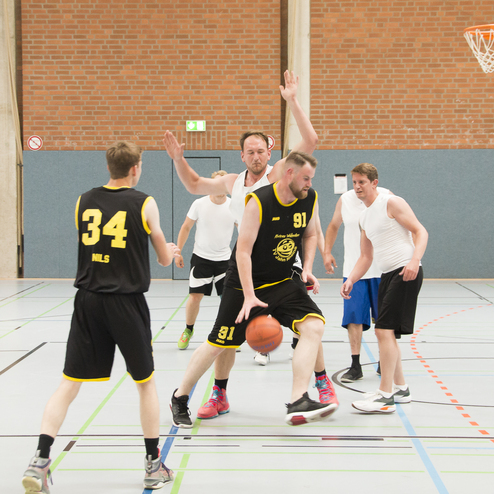 F&uuml;nf Basketballspieler in einem Spiel; zwei tragen Trikots, einer dribbelt den Ball, w&auml;hrend andere versuchen, ihn zu verteidigen.