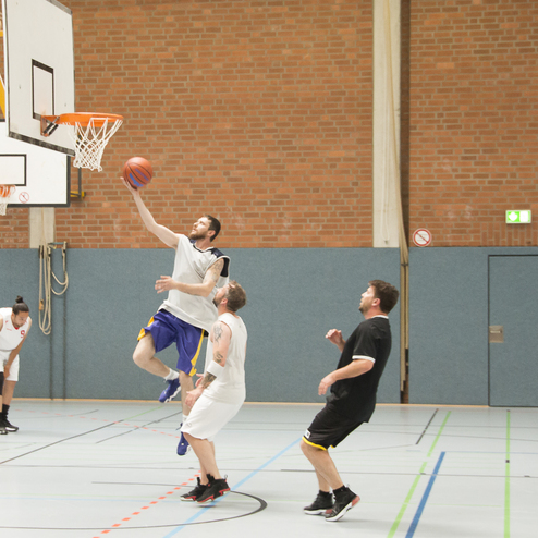 Basketballspieler springt zum Korb, w&auml;hrend andere Spieler in einem Sporthallen-Umfeld zuschauen.