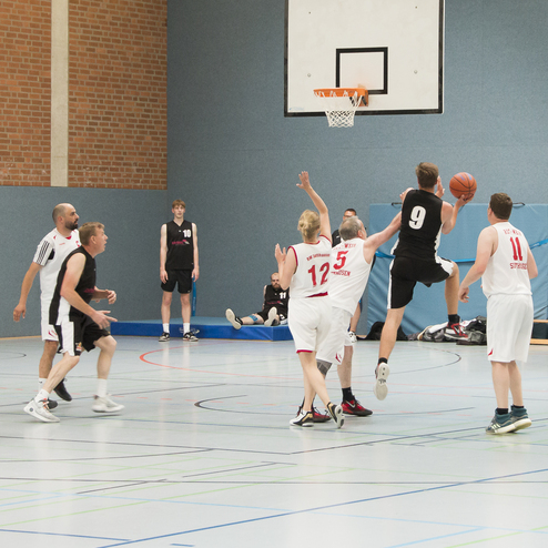 Zwei Basketballmannschaften spielen in einer Halle, ein Spieler angehoben mit Ball zum Korb.