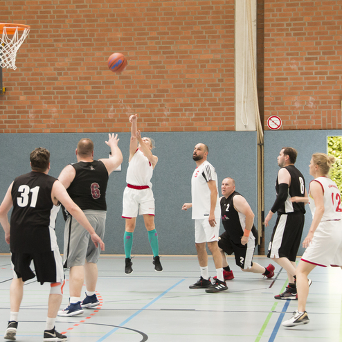 Ein Basketballspieler in wei&szlig;em Trikot springt zum Wurf, w&auml;hrend andere Spieler auf dem Spielfeld reagieren.