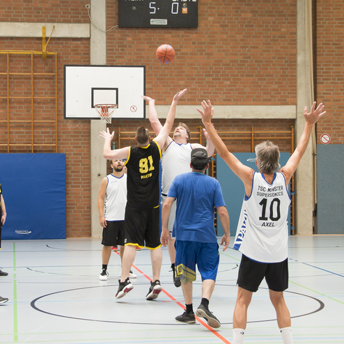 Basketballspiel in einer Halle, Spieler springen beim Sprungball, Korb im Hintergrund, Punkteanzeige zeigt 5:0.