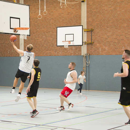 Spieler in Basketballtrikots beim Korbwurf in einer Sporthalle, im Hintergrund ein Kind und andere Spieler.