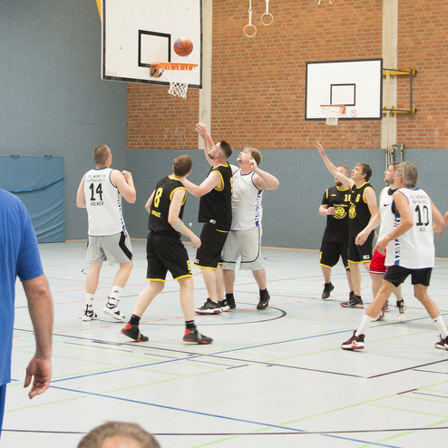 Basketballspiel in einer Halle, mehrere Spieler k&auml;mpfen um den Ball am Korb, Zuschauer im Hintergrund.