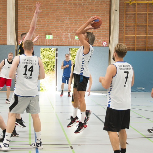 Basketballspieler werfen den Ball Richtung Korb w&auml;hrend eines Spiels in einer Sporthalle mit Sicht auf das Publikum.