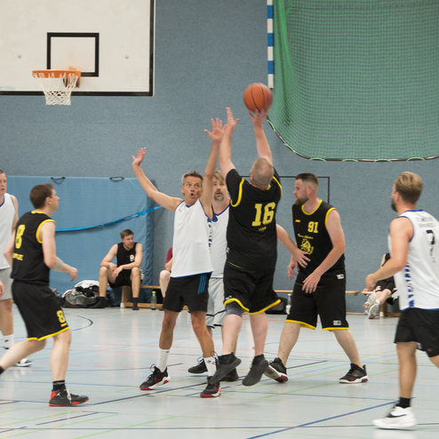 Zwei Basketballspieler k&auml;mpfen um den Ball, w&auml;hrend andere Spieler im Hintergrund auf dem Spielfeld aktiv sind.