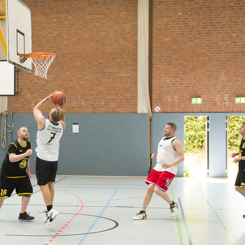 Spieler mit Trikot Nummer 7 wirft einen Basketball in einem Sporthallen-Spiel mit drei verteidigenden Spielern.