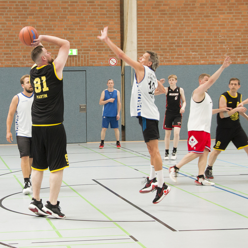 Spielszene in einer Sporthalle: Spieler in schwarzen und wei&szlig;en Trikots bei einem Basketballwurf und Abwehraktion.
