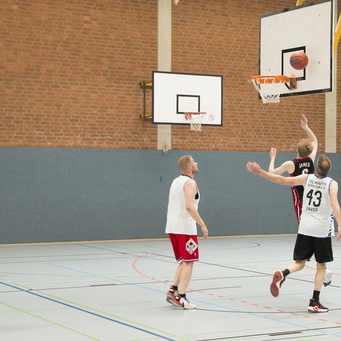 Vier Basketballspieler auf einem Feld; einer wirft den Ball auf den Korb, w&auml;hrend die anderen zuschauen.