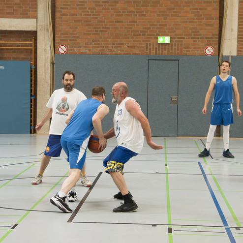 Sechs Spieler in Sportkleidung spielen Basketball in einer Halle, w&auml;hrend ein Spieler dribbelt und verteidigt wird.