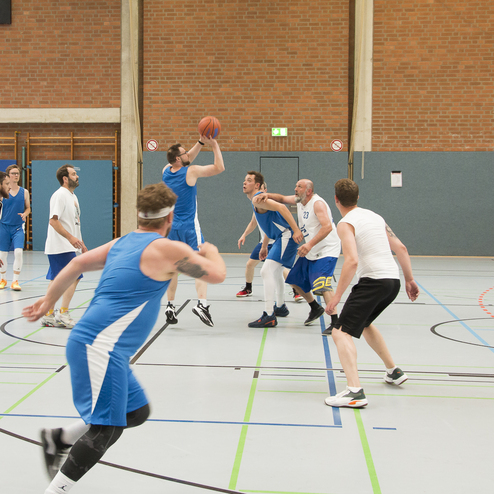 Szenenbild eines Basketballspiels mit Spielern in blauen und wei&szlig;en Trikots beim Korbwurf in einer Sporthalle.