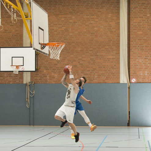 Basketballspieler im Sprung, der einen Korb wirft, w&auml;hrend ein anderer Spieler verteidigt. Hallenboden und Basketballkorb sichtbar.