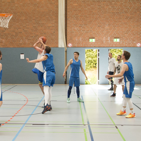 Spielszene in einer Sporthalle: Ein Spieler wirft den Basketball auf den Korb, w&auml;hrend andere Spieler reagieren.