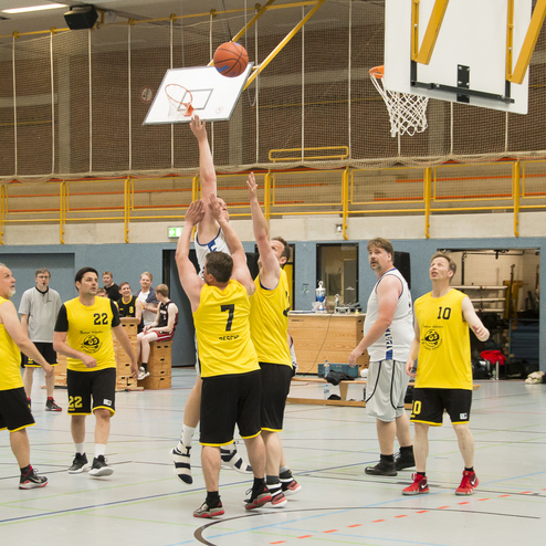 Mehrere Basketballspieler im Gelb und Wei&szlig; k&auml;mpfen um den Ball unter dem Korb in einer Sporthalle.