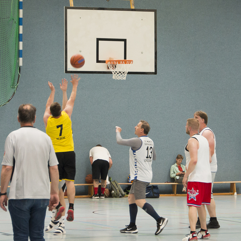 Zwei Basketballspieler werfen den Ball Richtung Korb, w&auml;hrend vier weitere Spieler im Hintergrund aktiv sind.