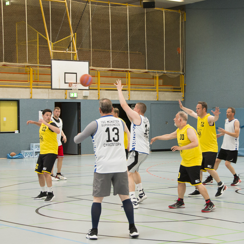 Zehn M&auml;nner spielen Basketball in einer Sporthalle, w&auml;hrend ein Spieler den Ball wirft.