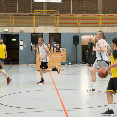 Zwei Basketballteams spielen in einer Sporthalle; Spieler in gelben und wei&szlig;en Trikots bewegen sich mit dem Ball.