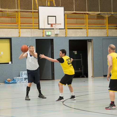Ein Basketballspieler in blauer Uniform wirft den Ball, w&auml;hrend ein Gegner in gelber Uniform ihn verteidigt.
