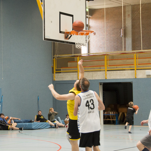 Zwei Basketballspieler greifen nach einem Ball, w&auml;hrend andere im Hintergrund auf einer Sportbank sitzen.