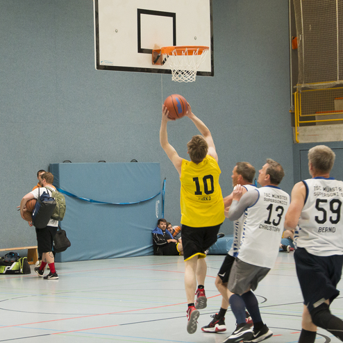Spieler in gelbem Trikot macht einen Wurf auf den Basketballkorb, w&auml;hrend andere Spieler in wei&szlig;en Trikots verteidigen.