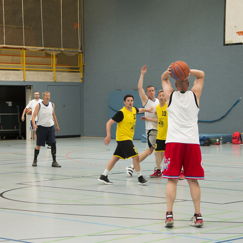 Spielszene in einer Halle: Ein Spieler in roten Shorts wirft einen Basketball, w&auml;hrend andere Spieler in gelben Trikots verteidigen.