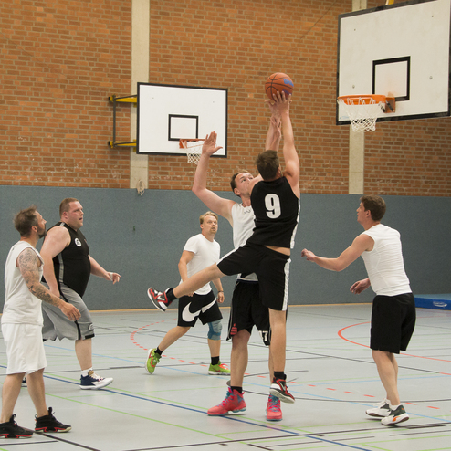 Basketballspiel in einer Halle: F&uuml;nf Spieler k&auml;mpfen um den Ball unter dem Korb, zwei Spieler springen hoch.
