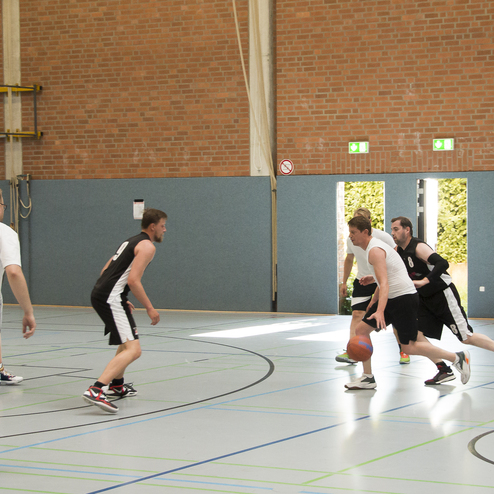 Sechs M&auml;nner spielen Basketball in einer Sporthalle, w&auml;hrend einer den Ball dribbelt und von einem Gegenspieler verfolgt wird.