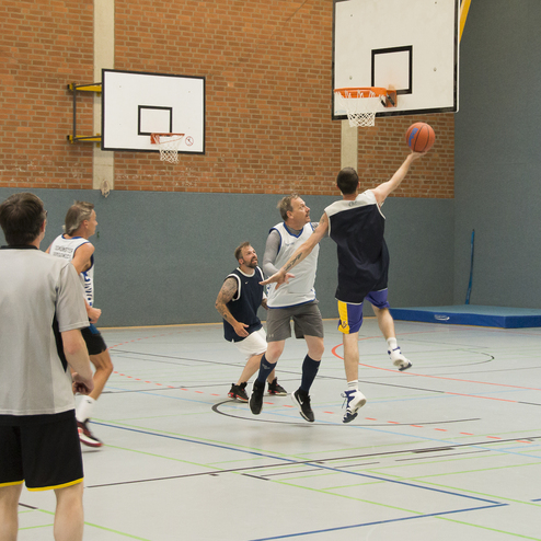 Basketballspiel in der Halle mit mehreren Spielern, einer springt zum Korb, w&auml;hrend andere verteidigen.