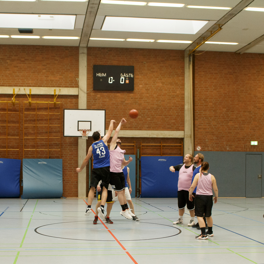 Zwei Basketballspieler springen zum Sprungball, w&auml;hrend vier weitere Spieler auf dem Feld positioniert sind.