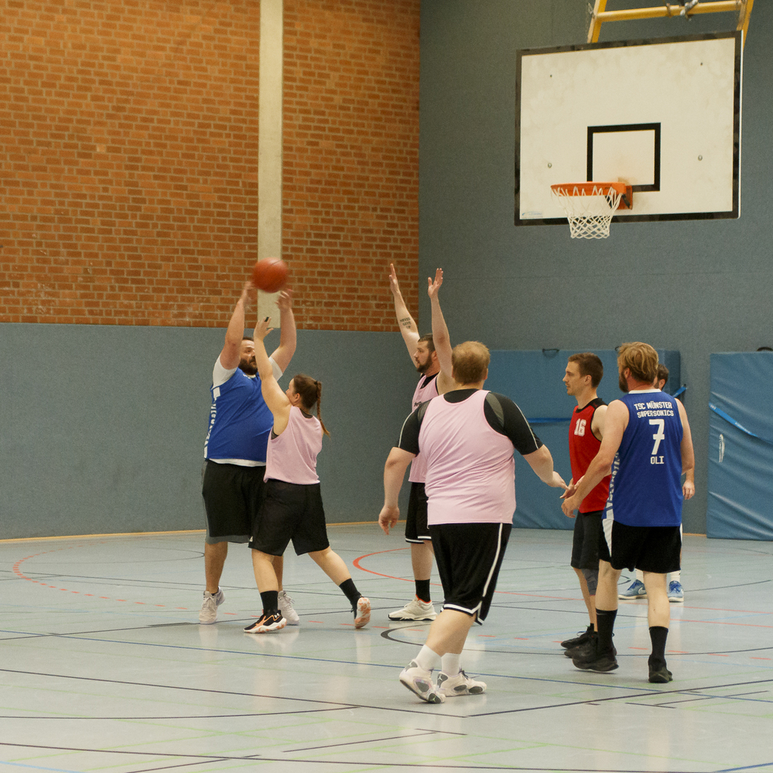 Zwei Basketballspieler werfen den Ball w&auml;hrend eines Spiels in einer Sporthalle, umgeben von weiteren Spielern.