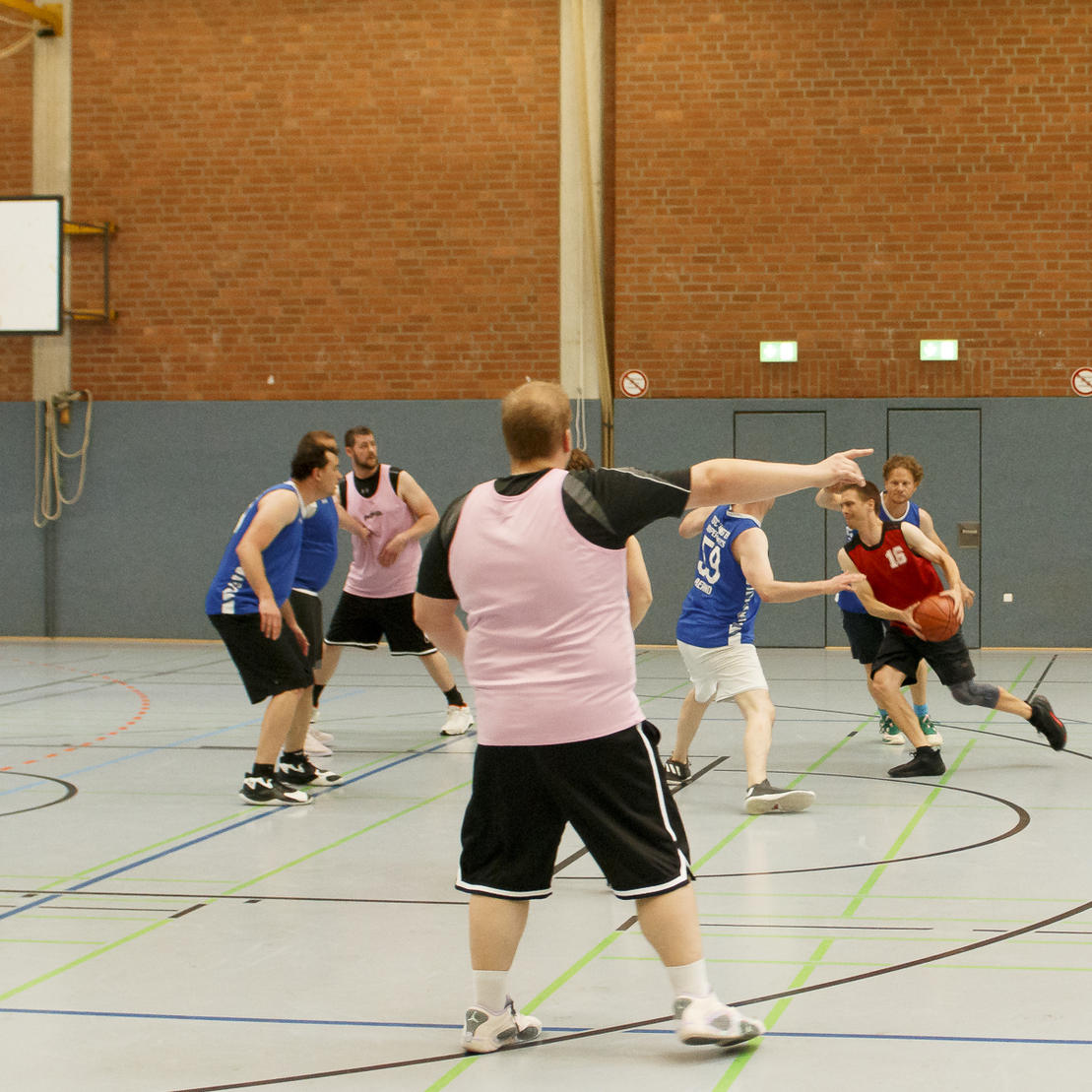 Basketballspiel in einer Halle mit Spielern in unterschiedlichen Trikots, die um den Ball anvisieren.