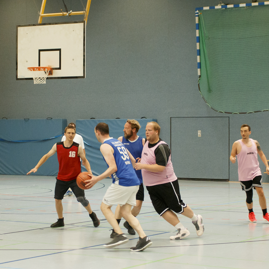 Siebene Spieler in Basketballtrikots stehen in der Sporthalle um den Ball, w&auml;hrend einer dabei ist, zu dribbeln.