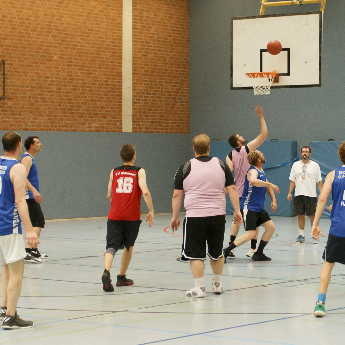 Basketballspiel in der Halle, Spieler werfen einen Ball auf den Korb, verschiedene Trikots in pink und blau.