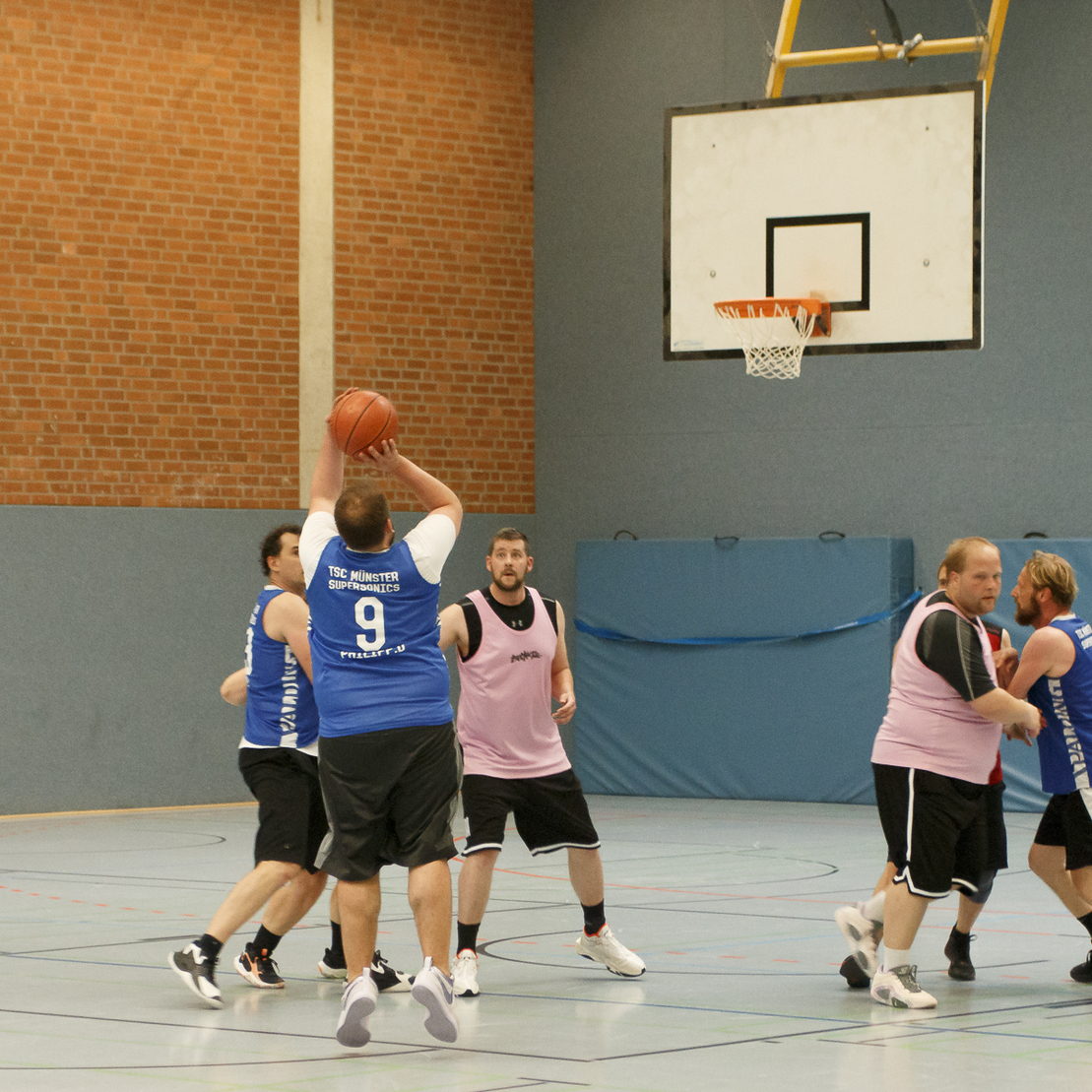 Ein Basketballspieler in Schwarz wirft den Ball zu einem Mitspieler w&auml;hrend eines Spiels in einer Sporthalle.