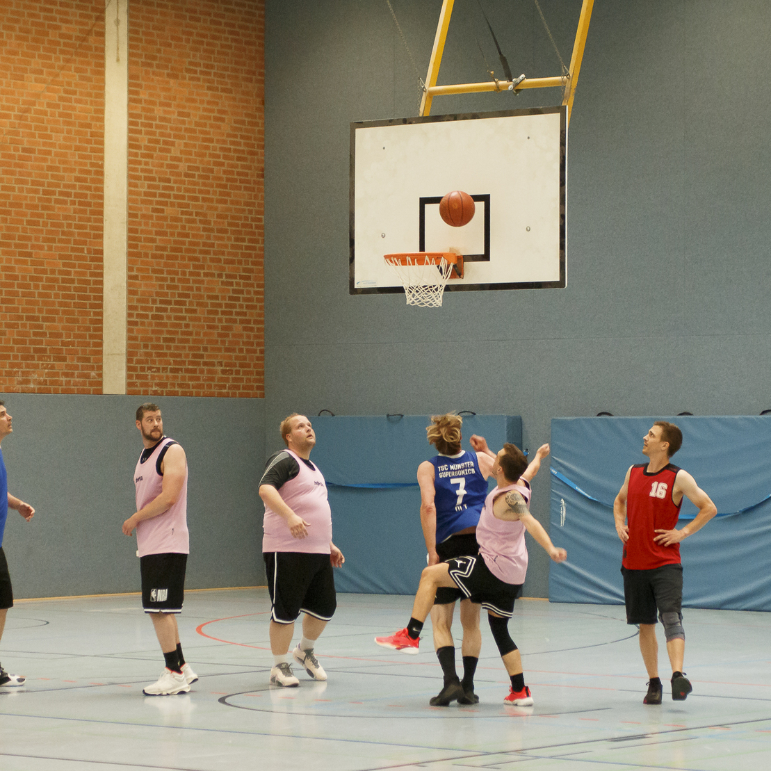 Spielszene in einer Halle: Basketballspieler werfen auf den Korb, einige springen, andere beobachten das Spiel.