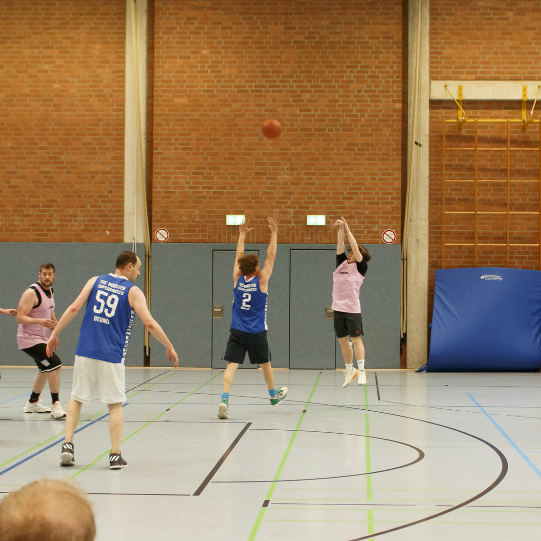 Basketballspiel in einer Halle, ein Spieler springt zum Wurf, w&auml;hrend andere in blauen und rosa Trikots positioniert sind.
