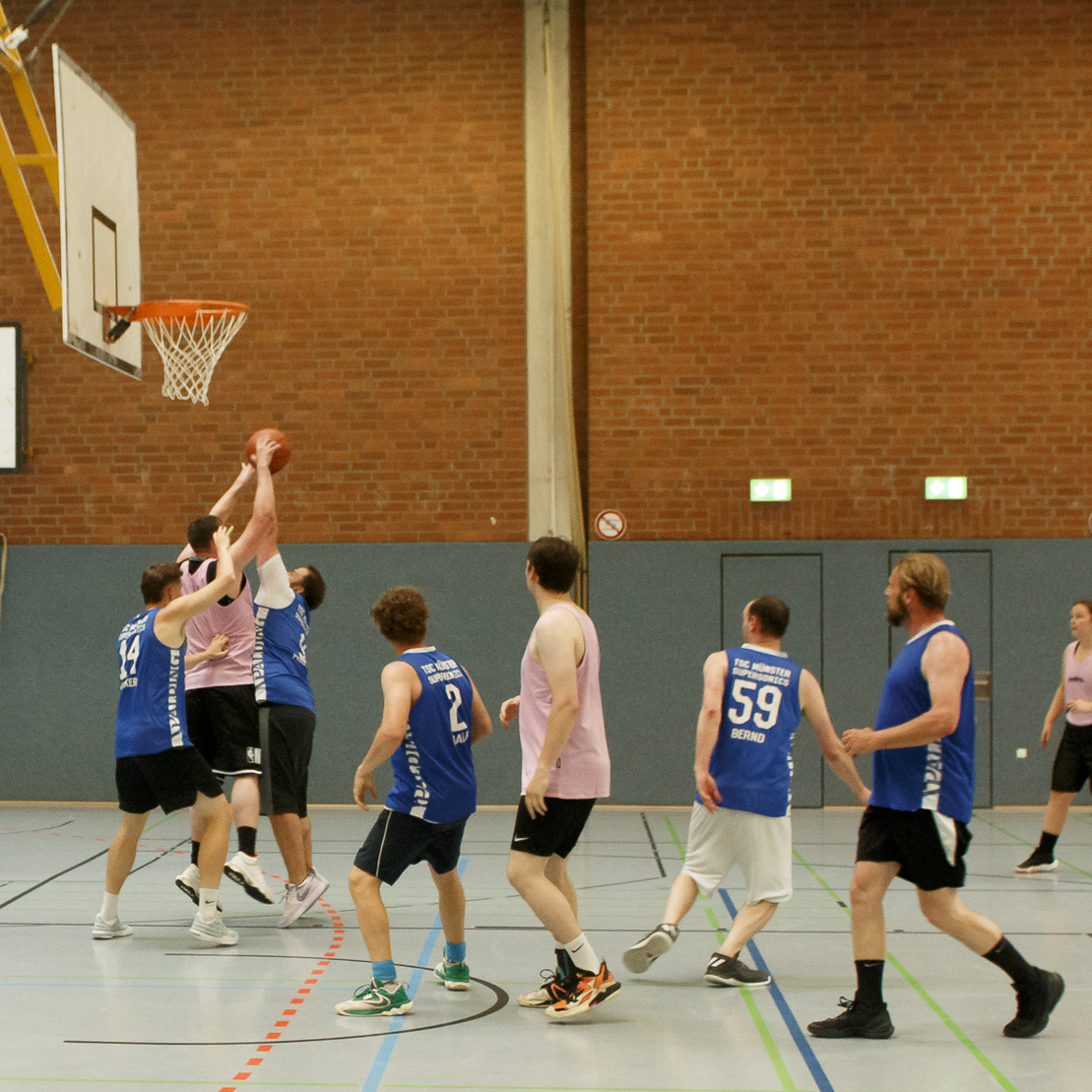 Basketballspiel in der Halle, mehrere Spieler in blauen und pinken Trikots k&auml;mpfen um den Ball beim Wurfkorb.
